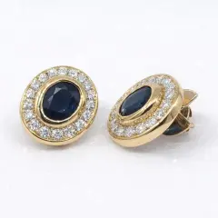 Pair of luxurious 18k yellow gold stud earrings featuring deep blue oval sapphires of 4.0ct totaling encircled by a halo of 32 sparkling round-cut diamonds of 1.28ct.