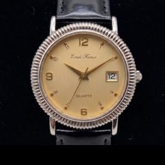 Emile France quartz watch. The watch features a champagne-coloured dial with gold-toned hands and a combination of Arabic numerals and dot indices. A small date window is positioned at the 3o'clock mark. The watch face is encased in a distinct silver-toned fluted bezel and is attached to a polished black leather strap.