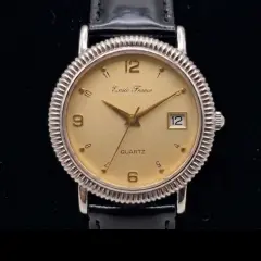 Emile France quartz watch. The watch features a champagne-coloured dial with gold-toned hands and a combination of Arabic numerals and dot indices. A small date window is positioned at the 3o'clock mark. The watch face is encased in a distinct silver-toned fluted bezel and is attached to a polished black leather strap.