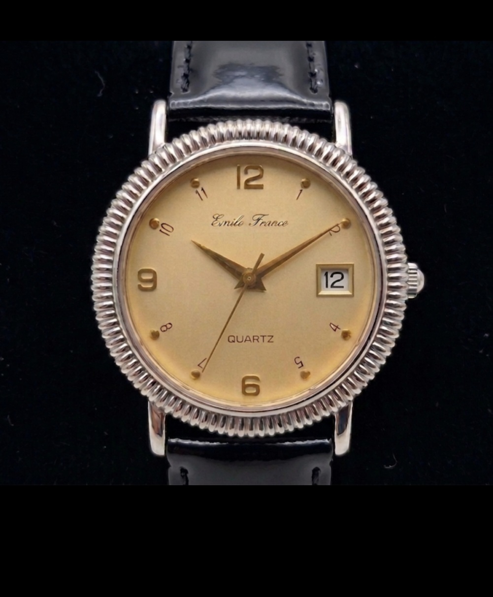 Emile France quartz watch. The watch features a champagne-coloured dial with gold-toned hands and a combination of Arabic numerals and dot indices. A small date window is positioned at the 3o'clock mark. The watch face is encased in a distinct silver-toned fluted bezel and is attached to a polished black leather strap.
