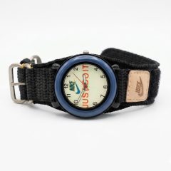 1780664039466 A Nike analog watch featuring a blue bezel and a black fabric strap. The watch face has a beige background with NIKE logo and the text "JUST DO IT" visible, and a small tan-coloured Nake brand patch is attached to the strap.